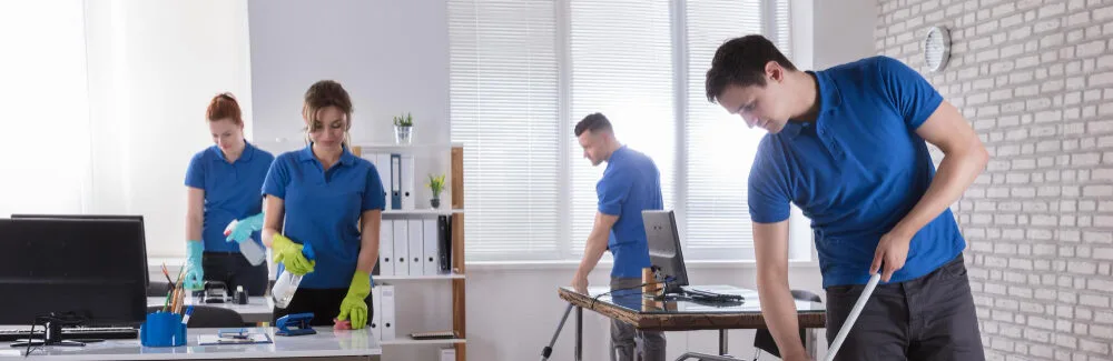 Cleaning professionals in blue uniforms actively tidying an office space, showcasing teamwork and attention to detail in a bright, modern environment.