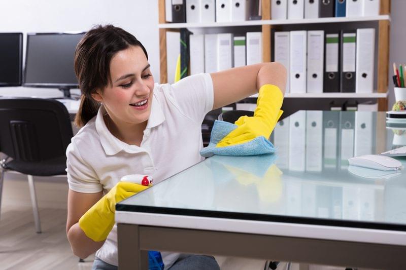 Close-up Of A Smiling Young Woman Cleaning The Glass Office Desk With Rag In Office